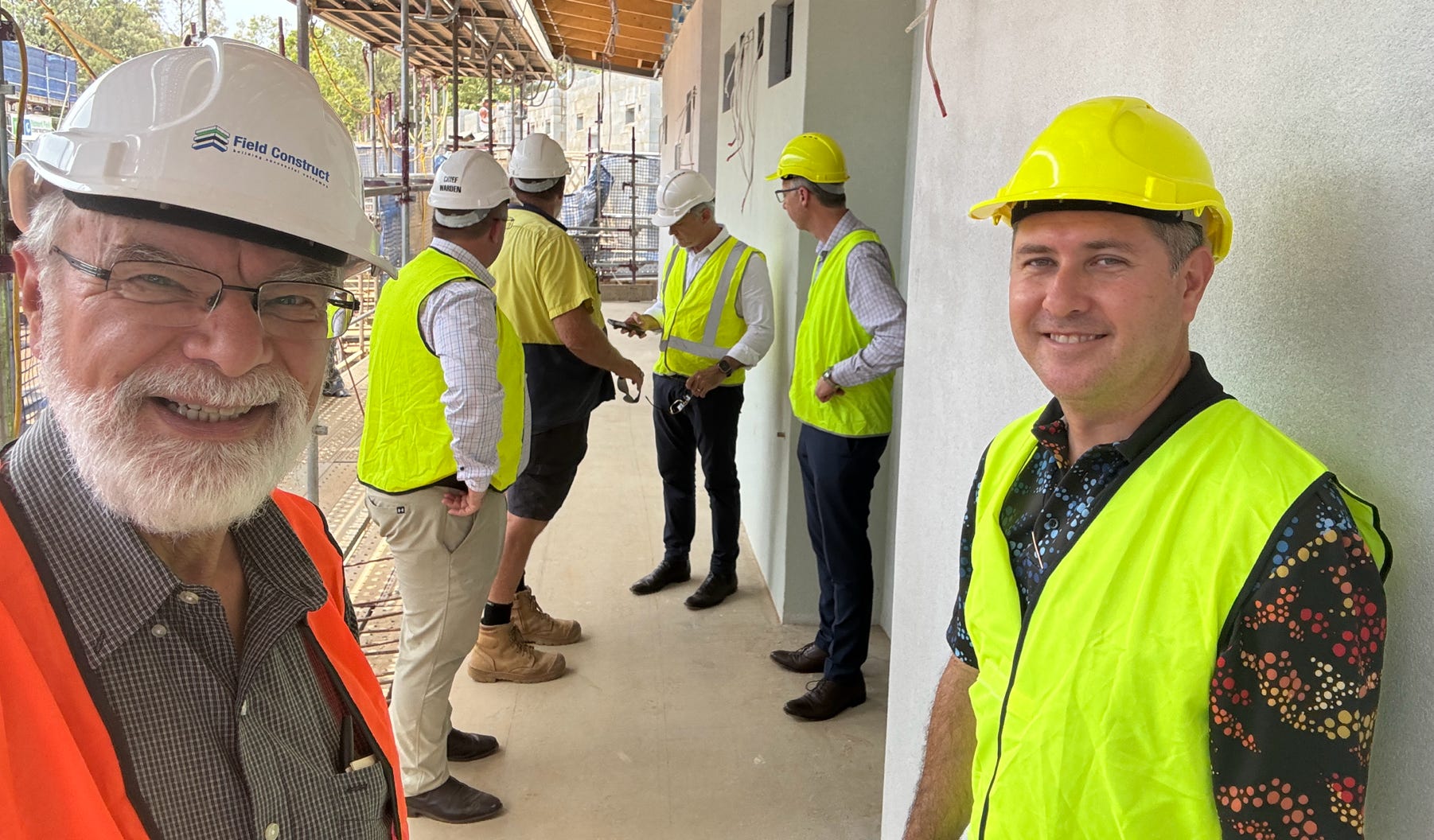 A group of workers with hard hats and hi-visibility clothing inspect construction work on a school campus.